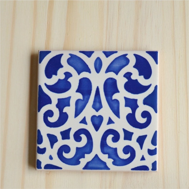 May include: A white ceramic tile with a blue and white intricate pattern. The pattern is a repeating design of swirls and curves.