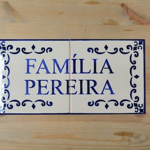 May include: Two white ceramic tiles with blue trim and decorative swirls. The tiles are joined together and display the text "FAMÍLIA PEREIRA" in blue lettering.