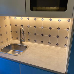 Op de afbeelding: Een kleine keukengootsteen met een chromen kraan en een lichtgrijs aanrechtblad. De achterwand is betegeld met witte tegels met een blauw-wit patroon. De kasten zijn blauw.