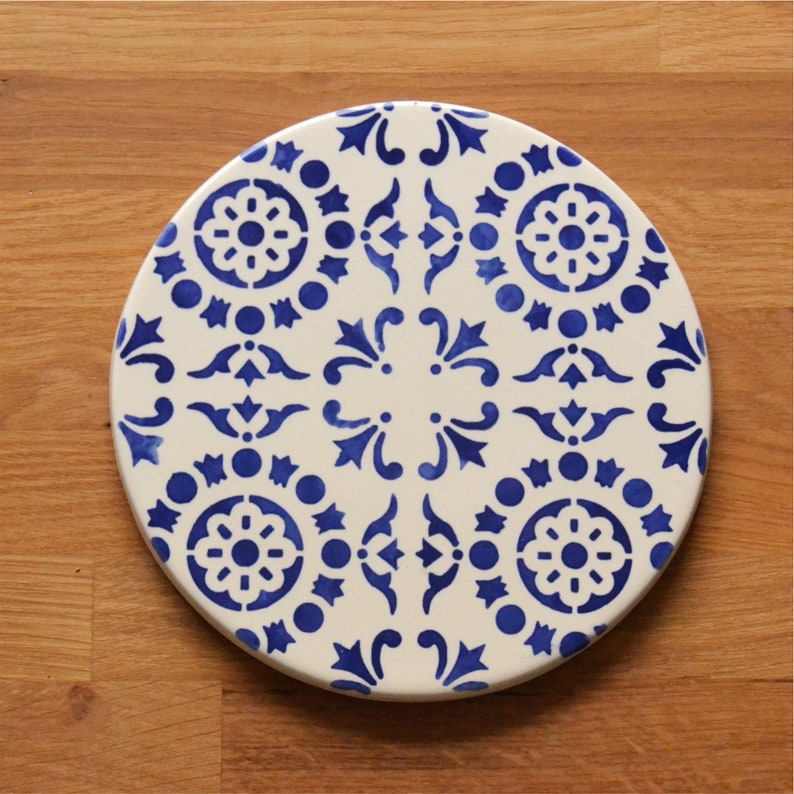 May include: A round ceramic trivet with a blue and white geometric pattern. The trivet features a repeating design of stylized flowers and leaves.