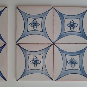 May include: Four white ceramic tiles with a blue and white floral design. Each tile features a stylised flower with four petals in the centre of a diamond shape.