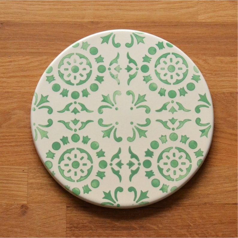 May include: A round ceramic coaster with a green and white geometric pattern. The coaster features a repeating design of stylized flowers and swirls.