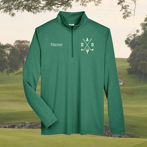 May include: A green long-sleeve golf shirt with a quarter-zip collar. The shirt features the word "Name" on the left chest and a white graphic with the letters "DAD" and golf clubs on the right chest. The shirt is made of a lightweight material.