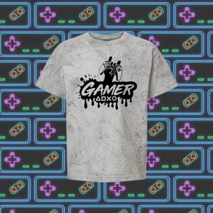 May include: A gray marble-patterned t-shirt featuring a black graphic of a video game controller and the word "GAMER" with dripping paint effect. The background is a pattern of neon-colored game controllers.