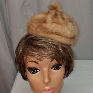 May include: A light brown fur hat, possibly mink, sits atop a mannequin head with short brown hair. The hat is a small, rounded style with a textured appearance. The mannequin has green eyes and a pink neck support.