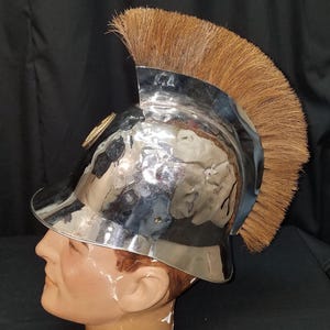 Könnte beinhalten: Glänzender, silberfarbener Helm mit einem braunen, bürstenartigen Kamm. Der Helm wird auf einem Schaufensterkopf präsentiert. Ein kleines, goldfarbenes Emblem befindet sich an der Seite. Der Hintergrund ist dunkel.