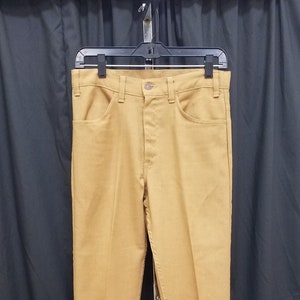 Puede incluir: Un par de pantalones de color marrón claro colgados de una percha negra sobre un fondo negro.