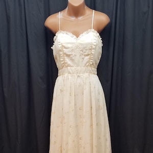 May include: A cream-colored, floor-length dress with spaghetti straps. The bodice features a sweetheart neckline with lace trim and embroidered detailing. The skirt has a tiered, flowing design with a subtle floral pattern.