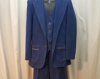 Può includere: Un completo in denim blu scuro composto da blazer, gilet e pantaloni. Il blazer presenta ampi revers, due tasche frontali con cuciture a contrasto e bottoni marroni. Il gilet abbinato ha quattro bottoni e i pantaloni hanno un taglio dritto.