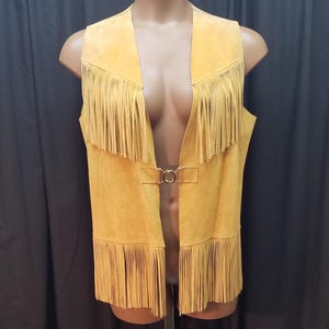 May include: A sleeveless yellow suede vest featuring long fringe detailing along the chest and the bottom hem. The garment is secured at the center front with a circular gold-toned metal clasp that connects two horizontal suede tabs.
