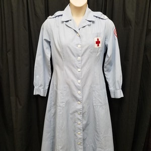 Puede incluir: Un vestido uniforme azul claro de manga larga con cuello blanco y cierre de botones. El vestido tiene un parche de cruz roja en el pecho izquierdo y dos bolsillos en la parte delantera.