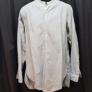 Può includere: Camicia azzurra a maniche lunghe con colletto alla coreana e abbottonatura frontale. La camicia presenta un sottile motivo a righe ed è esposta su un manichino. Le maniche hanno polsini con bottoni.