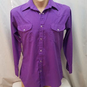 Puede incluir: Camisa morada lisa con botones, mangas largas, dos bolsillos en el pecho y cuello puntiagudo. La camisa se exhibe en un maniquí y tiene un estilo occidental clásico.