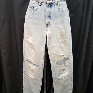May include: Light blue denim jeans with distressed details, including rips and patches. The jeans have a classic five-pocket design and a button and zipper closure. The jeans are displayed on a stand against a black backdrop.