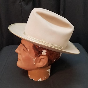 May include: A light beige fedora hat with a slightly curved brim and a decorative band. The hat is displayed on a mannequin head. The hat has a classic, timeless design, suitable for various occasions. The hat's color is neutral, making it versatile for different outfits.