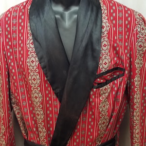 May include: A red robe featuring a repeating pattern of gold and white geometric motifs and vertical stripes. The garment includes a solid black shawl collar, a black waist tie, black pocket trim, and black sleeve cuffs, displayed on a mannequin.