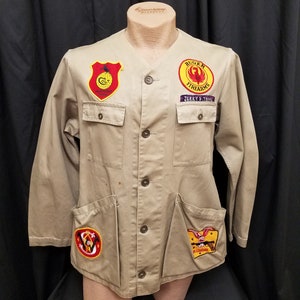 May include: A tan work jacket with multiple patches sewn on. The patches are yellow, red, and black with various logos, including "Colt", "Ruger Firearms", and "Jerry E. Trice".
