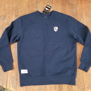 May include: Navy blue sweatshirt with a crew neck and long sleeves. A white and silver emblem is on the upper left chest. A small tag is attached to the collar. A white tag is on the lower left side.