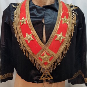 Könnte beinhalten: Schwarze Satinjacke mit rotem und goldenem Zierkragen. Der Kragen hat goldene Sterne und Fransen. Die Jacke hat goldene Verzierungen an den Ärmeln und ein goldenes Emblem am unteren Rand.