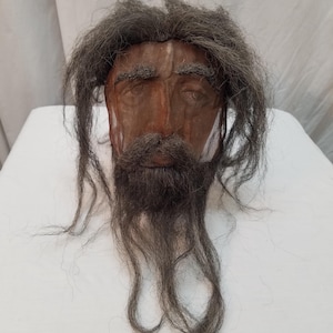 May include: A sculpted head with a brown face, dark gray hair, and a long beard. The face has detailed features, including a prominent nose and deep-set eyes. The hair and beard appear to be made of a dark, textured material.