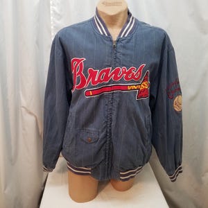 Op de afbeelding: Blauw baseballjack met witte krijtstrepen en het woord "Braves" in rode letters over de borst. De jas heeft een ritssluiting, twee voorzakken en een patch op de mouw.