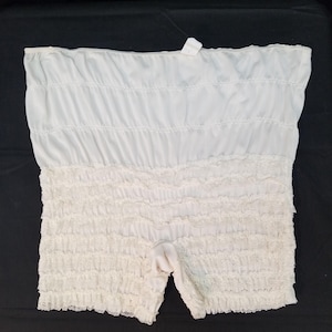 May include: White ruffled shorts with a high waist. The top portion is a smooth, gathered fabric, while the bottom features a textured, ruffled design. A small tag is attached at the top. The shorts are made of a soft, lightweight material.