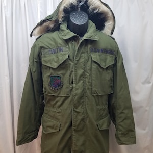 以下が含まれることがあります： オリーブグリーンのUS Air Forceパーカ、毛皮の裏地付きフード。 ジャケットの左胸ポケットの上には「MARTIN」、右胸ポケットの上には「U.S. AIR FORCE」と書かれています。 左胸にはカラフルなエンブレムのパッチが付いています。 ジャケットには複数のポケットとドローストリングウエストが付いています。