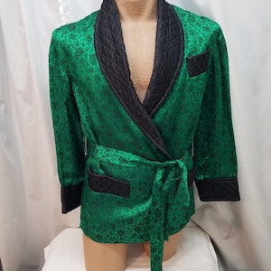 Op de afbeelding: Een groen smokingjack met patroon, zwarte gewatteerde revers, zakafwerking en manchetten. De jas heeft een bijpassende strikriem en wordt op een mannequin gepresenteerd. De stof heeft een subtiel bloemmotief.