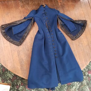 May include: A long, navy blue coat with a high collar and a row of black buttons down the front. The coat features unique flared sleeves trimmed with a dark, decorative border. The coat is displayed on a wooden surface.