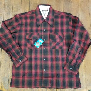 May include: A red and black plaid long-sleeve shirt with a button-down collar and two chest pockets. The shirt has a tag attached to the pocket.