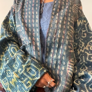 Könnte beinhalten: Blau-weiß gemusterte Jacke mit gesteppter Textur. Die Jacke hat eine lockere Passform und weite Ärmel. Das Muster umfasst geometrische Formen und vertikale Streifen. Die Jacke wird über einem blauen Oberteil getragen.