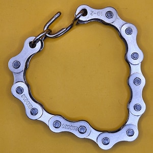 May include: A silver bicycle chain bracelet with a hook clasp. The bracelet is made of interlocking metal links, with the brand name "KMC" visible on some links. The bracelet is displayed against a yellow background.
