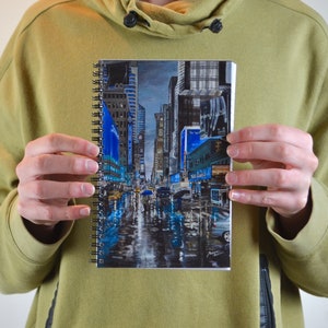 May include: A spiral bound notebook with a colorful painting of a rainy city street scene. The painting features tall buildings, streetlights, and people walking with umbrellas.