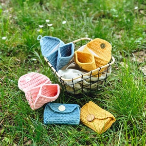 Könnte beinhalten: Handgefertigte Häkeltaschen und Geldbörsen in Blau, Rosa und Gelb. Einige befinden sich in einem Drahtkorb, andere auf grünem Gras. Jedes Stück hat einen Knopfverschluss.