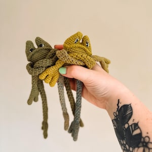 Könnte beinhalten: Zwei handgefertigte Froschspielzeuge aus Häkelarbeit in Grün- und Gelbtönen werden hochgehalten. Die Frösche haben lange Beine und kleine, detaillierte Merkmale. Die Spielzeuge sind weich und scheinen aus Garn hergestellt zu sein.