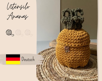 May include: A handmade, yellow-gold crochet pineapple-shaped basket with a green crown. The basket has a lid and sits on a woven mat. The image includes the text "Utensilo Ananas" and a German flag with the word "Deutsch".