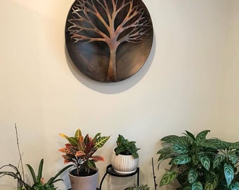 May include: A circular metal wall art piece featuring a tree silhouette with copper and dark brown tones, displayed above a collection of various potted houseplants arranged on multiple black metal plant stands of differing heights.