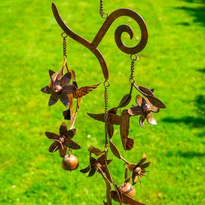 May include: A bronze-colored metal wind chime featuring hummingbirds, flowers, and small round accents. The wind chime is suspended from a decorative hook and hangs against a backdrop of green grass.