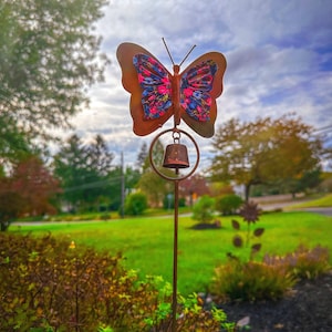 May include: A metal garden stake featuring a butterfly design with a bell. The butterfly has a rusty brown outline and a colorful floral pattern in pink, blue, and white. The bell hangs below the butterfly, and the stake is placed in a garden.