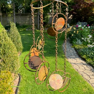 May include: A copper wind chime with multiple circular and teardrop-shaped metal pieces hanging from chains. The chime is suspended from a curved metal rod.