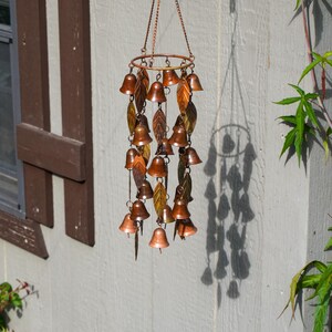 Puede incluir: Un carill&oacute;n de viento colgante con un marco circular de metal en tono cobre, que presenta m&uacute;ltiples adornos peque&ntilde;os en forma de campana y detalles met&aacute;licos en forma de hoja suspendidos por cadenas finas, con un acabado met&aacute;lico c&aacute;lido.