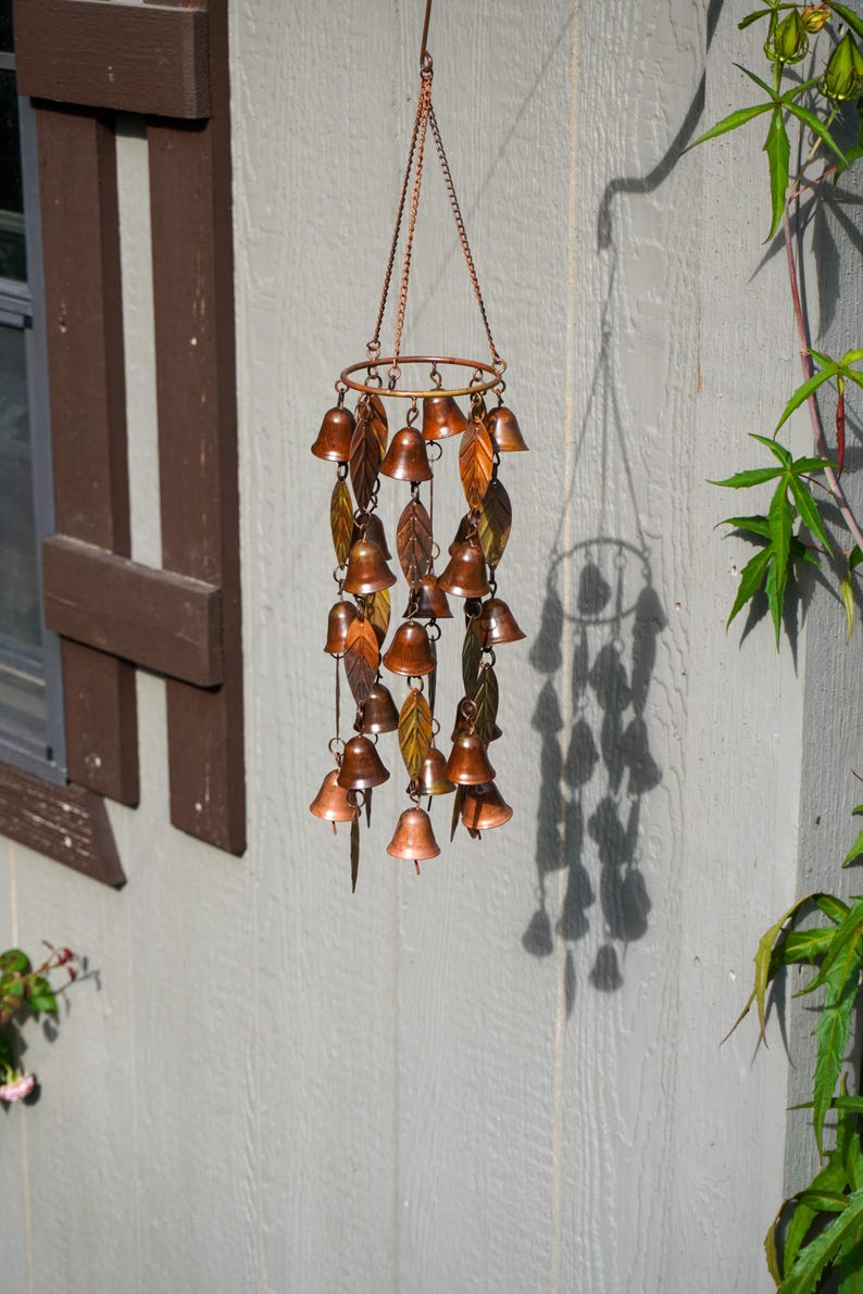 Puede incluir: Un carill&oacute;n de viento colgante con un marco circular de metal en tono cobre, que presenta m&uacute;ltiples adornos peque&ntilde;os en forma de campana y detalles met&aacute;licos en forma de hoja suspendidos por cadenas finas, con un acabado met&aacute;lico c&aacute;lido.