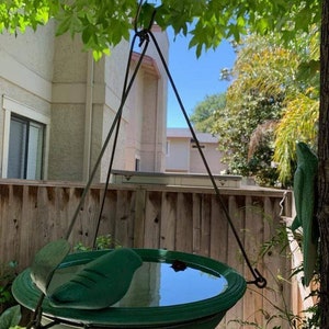 Puede incluir: Un bebedero para p&aacute;jaros verde con un dise&ntilde;o de hoja cuelga de un gancho de metal. El bebedero est&aacute; lleno de agua y refleja el cielo azul.