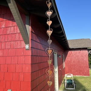 May include: A vertical rain chain made of interconnected copper-colored metal hearts hanging from the edge of a roof. The chain consists of ten heart-shaped links of varying sizes, suspended in a single line against a red shingled wall.