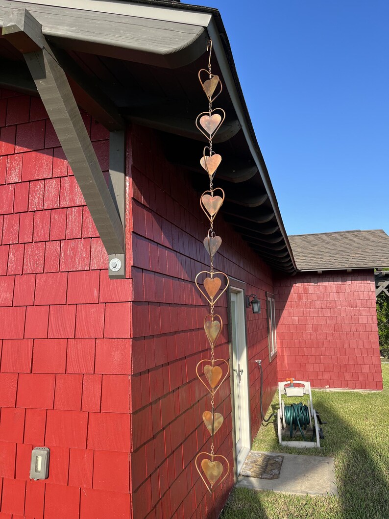 May include: A vertical rain chain made of interconnected copper-colored metal hearts hanging from the edge of a roof. The chain consists of ten heart-shaped links of varying sizes, suspended in a single line against a red shingled wall.