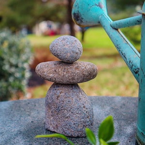 May include: A stack of three gray stones balanced on top of each other. The stones are smooth and round, and they are arranged in a pyramid shape. The stones are sitting on a gray surface, and there is a blue watering can in the background.