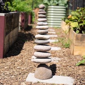 May include: A balanced stack of smooth, gray stones in a garden setting. The stones are arranged in a tower, with a large base stone and smaller stones stacked upwards. The background includes wooden planters and greenery.