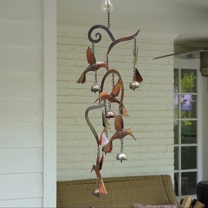 May include: A metal wind chime featuring multiple hummingbirds with copper and brown tones. The wind chime has a curved metal design with bells hanging from each hummingbird.
