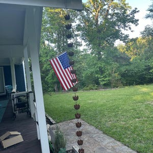 May include: A brown metal rain chain hangs from the gutter of a house. The chain is made up of a series of connected cups, each shaped like a flower. An American flag is hanging on the side of the house.
