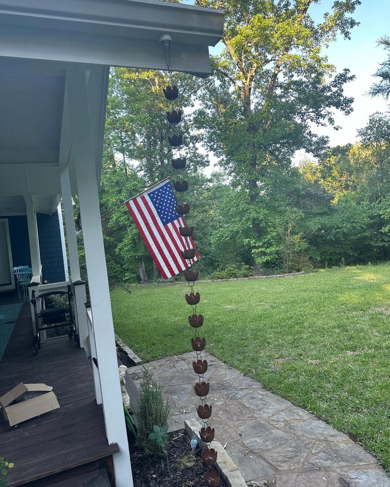 May include: A brown metal rain chain hangs from the gutter of a house. The chain is made up of a series of connected cups, each shaped like a flower. An American flag is hanging on the side of the house.